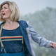 Jodie Whittaker's 13th Doctor rests against a stone column with her sonic screwdriver in hand, glancing at an unseen threat in the sky.